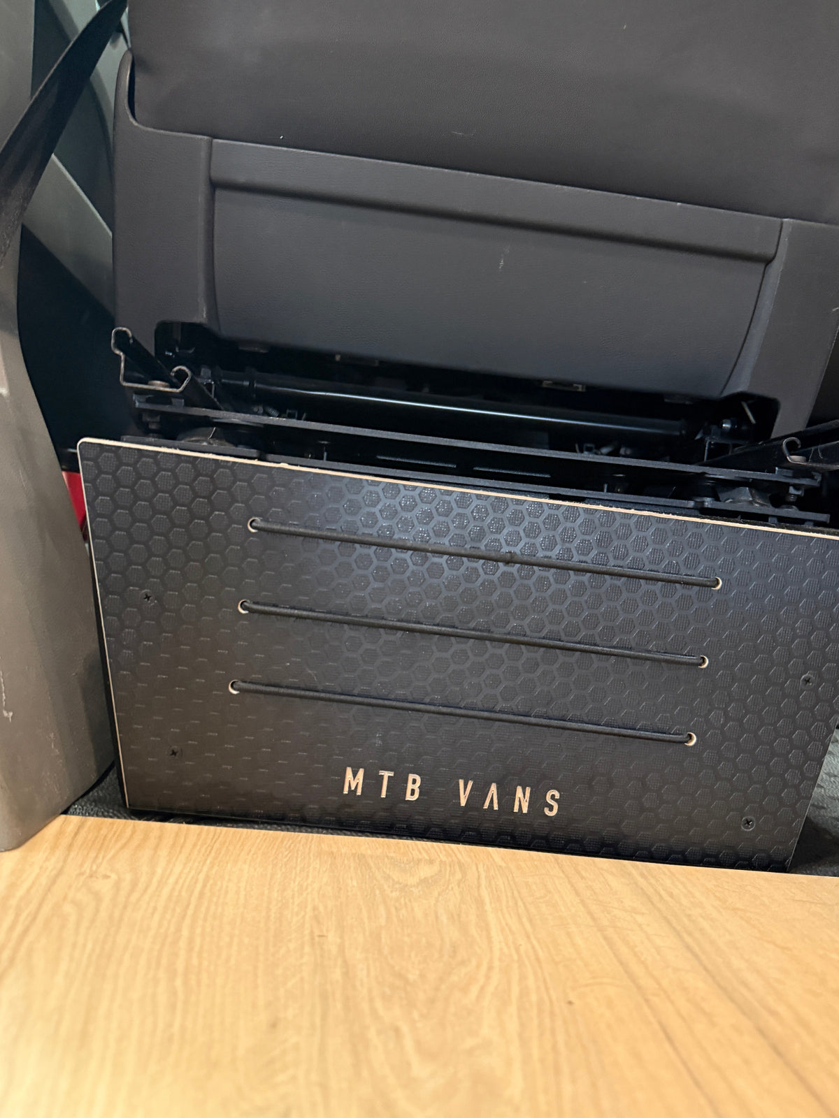 A black MTB Vans storage box with a honeycomb pattern and three horizontal handles, shown installed under a gray vehicle seat in a vehicle interior.